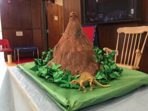 Picture of a brown paper mache volcano with green bushes and a dinosaur skeleton