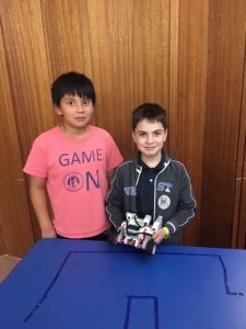 A boy in a salmon colored shirt stands next to a shorter boy in a blue jacket and shirt. The boy in blue rests an EV3 robot on a blue table. Both boys are smiling.