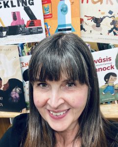A dark-haired smiling woman sits in front of a book display of picture books