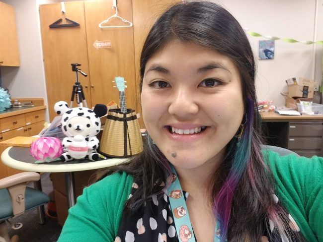 Melody Leung posing in front of storytime materials including a stuffed animal, ukelele, and audio equipment.