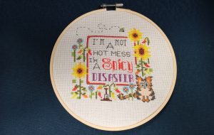 A cross stitch reads "I'm not a hot mess I'm a spicy disaster" surrounded by flowers and pepper plants.