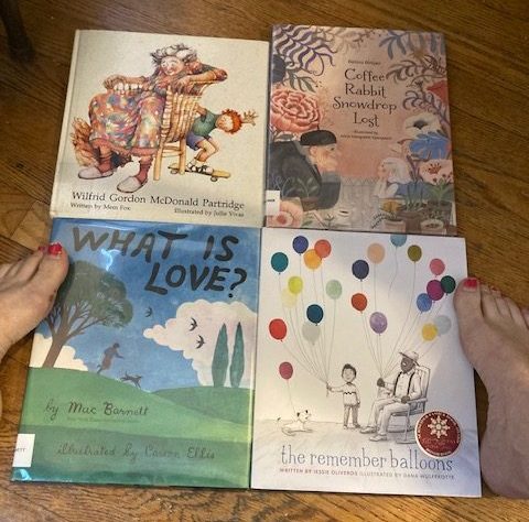 4 picture books that deal with children's relationship with people who have dementia. From left, first row: Wilfred Gordon McDonald Partridge; Coffee Rabbit Snowdrop Lost; 2nd row: What is Love; The Remember Baloons. Author's feet with ragged pink nail polish show up on either side of the books.