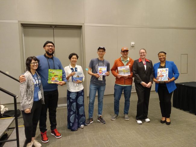 Moderate professional engagement: A group of authors poses with their books after a panel discussion. From left to right: illustrator Eliza Kinkz, author Jesus Trejo, author Rosa Chang, author Greg Pizzoli, author Aaron Becker, facilitator Jaime Eastman, and illustrator Erika Lynne Jones.