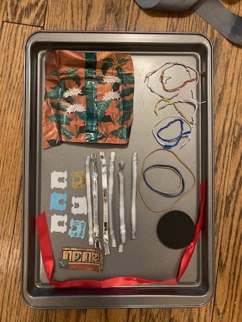 A cookie tray with a piece of flat plastic covered with flowers; some bread ties, arranged; some rubber bands, arranged; a red ribbon; a row of coffee wrapper ties; a row of bread tie discs, arranged; a piece of plastic with the number 1912.