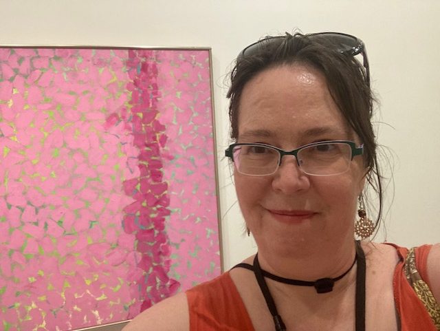 A painting of Alma Thomas that has light pink and darker pink stripes; a woman in an orange dress stands next to it. This is a selfie, so you only sese part of the painting and part of the woman.