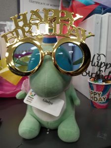 A green dinosaur in a birthday hat wears a pair of Happy Birthday sunglasses