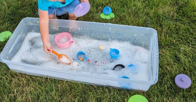 Bubble Tea Party Sensory Bin : long bin filled with bubble soap, tea set parts, pom-poms, and plastic spoons.