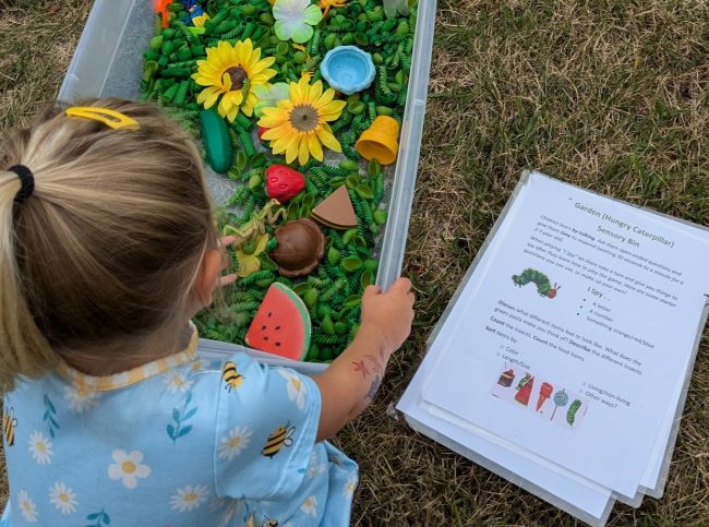 Very Hungry Caterpillar Sensory Bin; box filled with green pasta shapes, yellow flowers, ice cream, watermelon slice, strawberry and other items.
