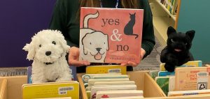 A white dog puppet sits with a black cat puppet on top of a shelf of books. Between them is a picture book titled Yes & No with a picture of a white dog and a black cat on the cover.