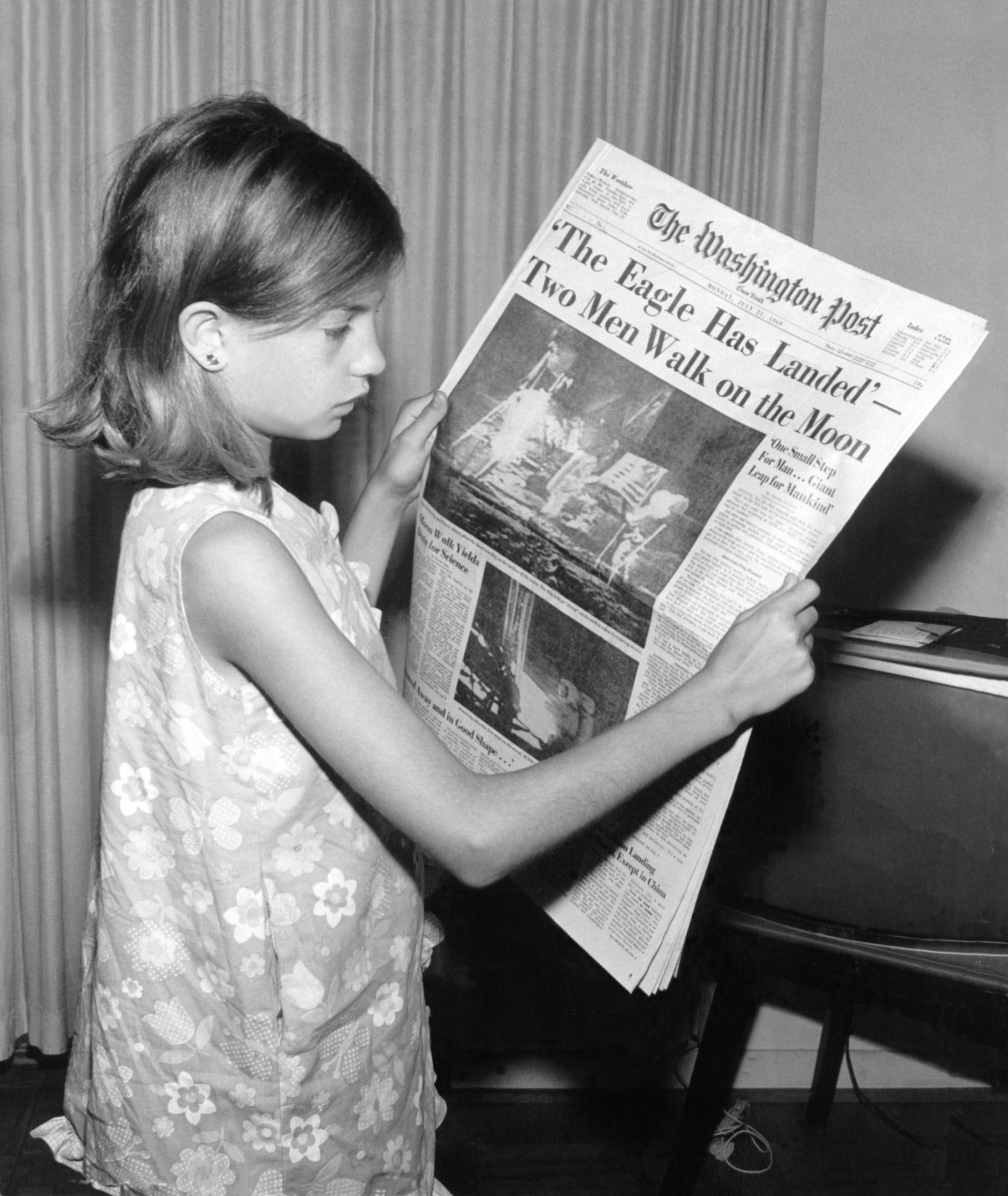 A young girl wearing a dress holds a Washington Post newspaper with the headline "'The Eagle Has Landed' - Two Men Walk On The Moon." A small TV is on her right and she is standing in front of a curtain. The photo is in black and white.
