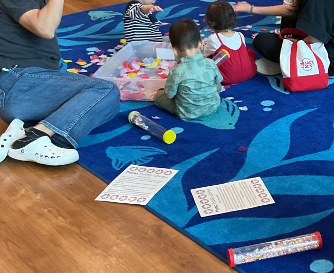 A  group of children sit on a blue rug. A plastic bin full of multicolored flower petals is in the center, with one child dropping petals into the bin. Another child holds a sensory tube.