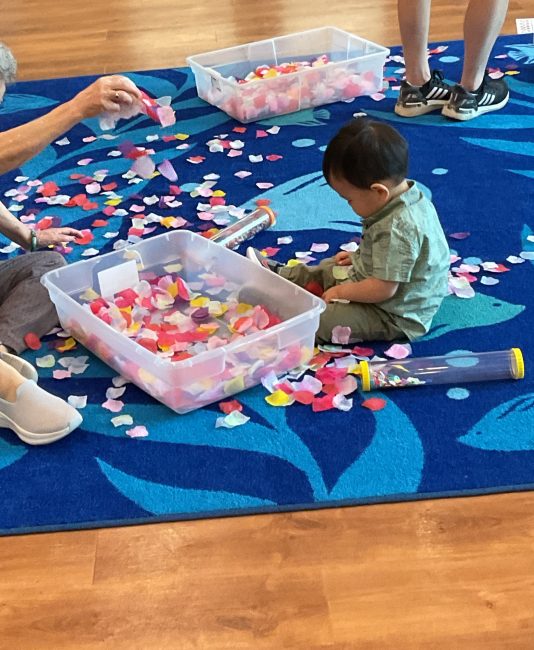 A dark haired child sits on a blue rug next to a plastic tub filled with multicolored flower petals. He holds an assortment of flower petals in his lap.