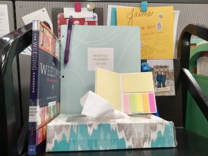 A black book cart shelf shows a weeding manual, a box of tissues, a calendar, a pad of sticky notes, and a Stitch pen.