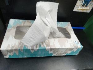 A blue and gray box of tissues sits on a desk with a single white tissue sticking out.