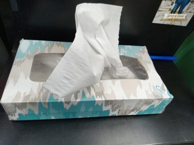 A blue and gray box of tissues sits on a desk with a single white tissue sticking out.