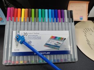 A box of pens sits on a desk, arranged in a rainbow from pink to black.