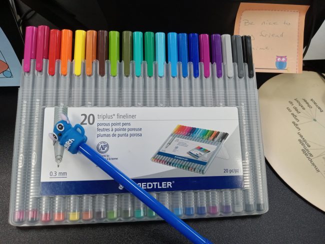 A box of pens sits on a desk, arranged in a rainbow from pink to black.