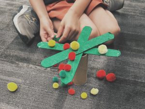 A child sits on a floor using a paper towel tube and craft sticks to create a tree. There are multi-colored pom poms on the tree and on the floor.
