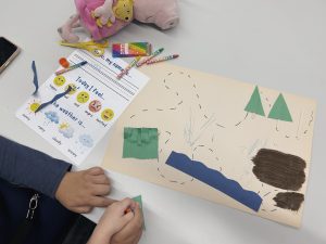 A family sitting at a table working on a craft. There is a sheet of paper next to the craft with questions about feelings and the weather, and crayons are scattered on top of it.