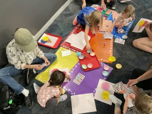 A group of children work on a craft using cupcake liners, glue sticks, and stickers. Craft supplies are displayed on colored mats on the floor with children all around.