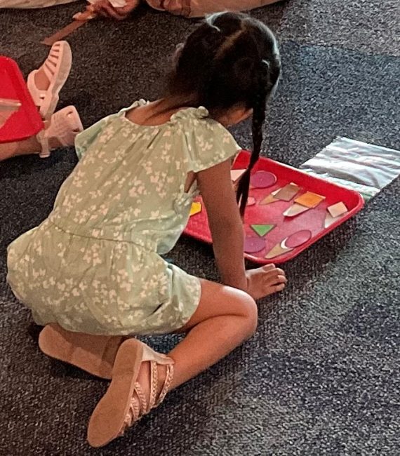 A child kneels on the floor with a red tray. On the tray are ice cream cones with different shapes for the scoop. She practices matching the shape to the right cone.