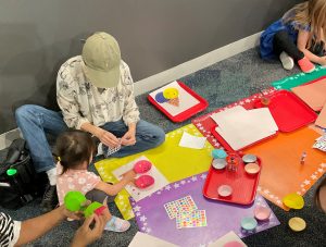 A caregiver and child work together on the floor to create an ice cream cone from cupcake liners and craft supplies.