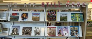 Photo of a library book display with block text on top that says, "We are still here."