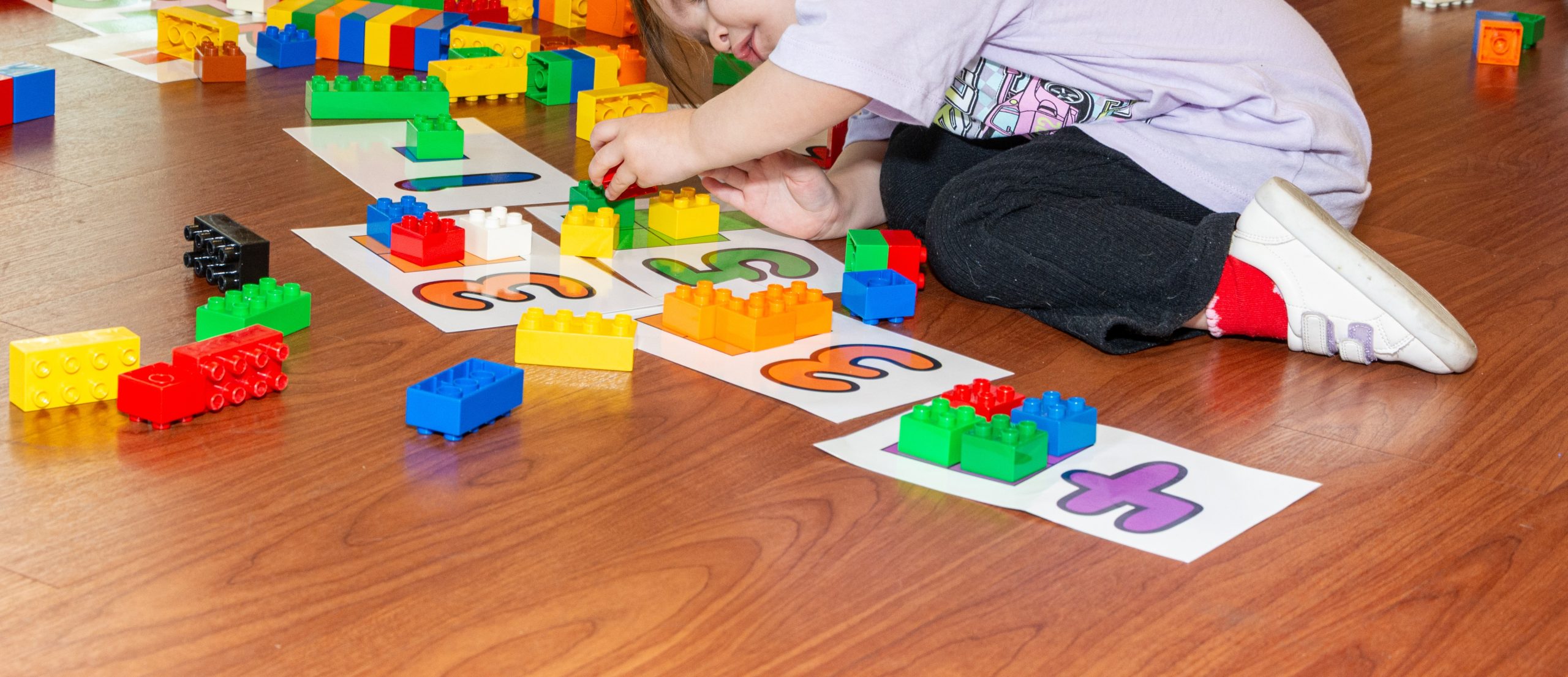 Colorful Duplo blocks are scattered on floor on top of white pieces of paper with large, full-page numbers. A child lies on the floor playing with blocks with only a leg and arm visible.