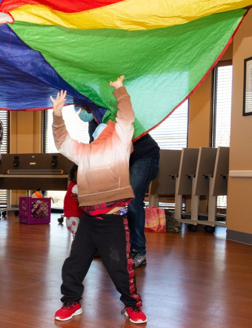 A child wearing black pants and a gray hoodie lifts their arms overhead with head tilted back to view a rainbow parachute overhead.