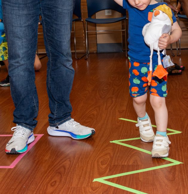 A child wearing a blue tshirt and blue shorts with green and orange apples, carrying a stuffed bird, walks along a green zig-zag line taped on the floor. To the left, a pair of adult legs in blue jeans appear.