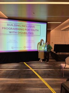 Photo of two female-presenting individuals standing to the right of a screen that reads, "Building Inclusive Programming for Youth with Disabilities"