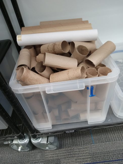 A plastic bin holds multiple toilet paper tubes and paper towel rolls.