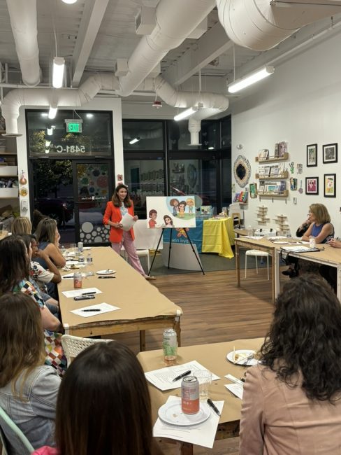 A photo of of a classroom, with tables in a "U" shape facing a white woman in a red blazer presenting images of families on a white background.