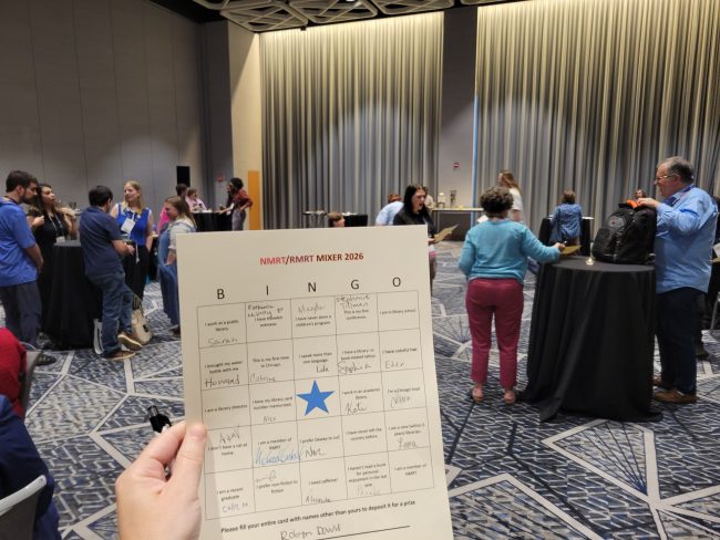 BINGO game card, with some squares already filled in, held in front of the reception where New Member Round Table and Retired Member Round Table ALA members were networking.