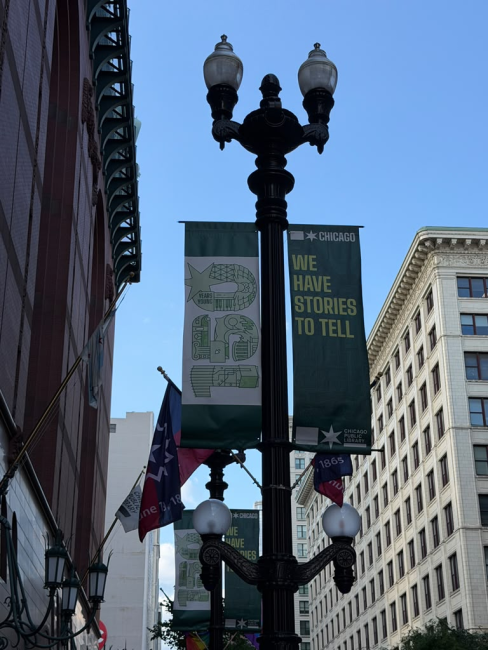 Sign hanging from a street lamp: "150 - CHICAGO - We have stories to tell" in front of the Chicago Public Library