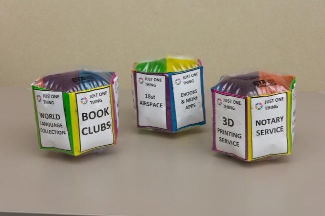 Three activity cubes sit side by side. Inside each insert is a card that reads Just One Thing and text about library services like Book Clubs or 3D printing.