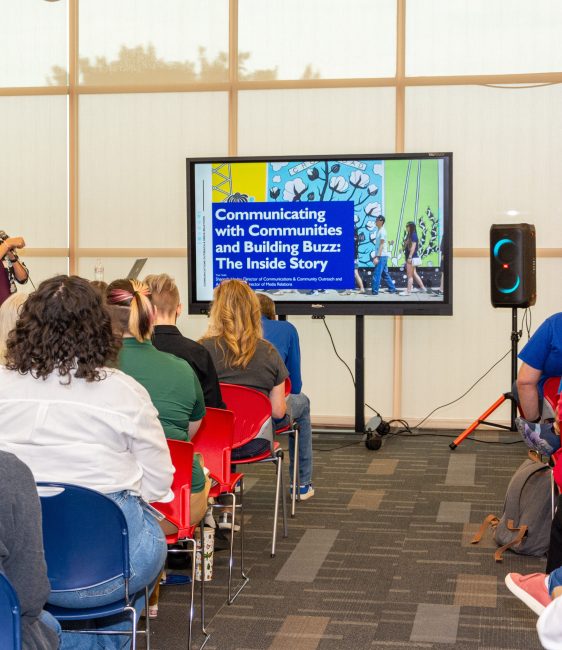 A group of adults sit facing a screen. The words Communicating with Communities and Building Buzz: The Inside Story appear on a presentation on the screen.